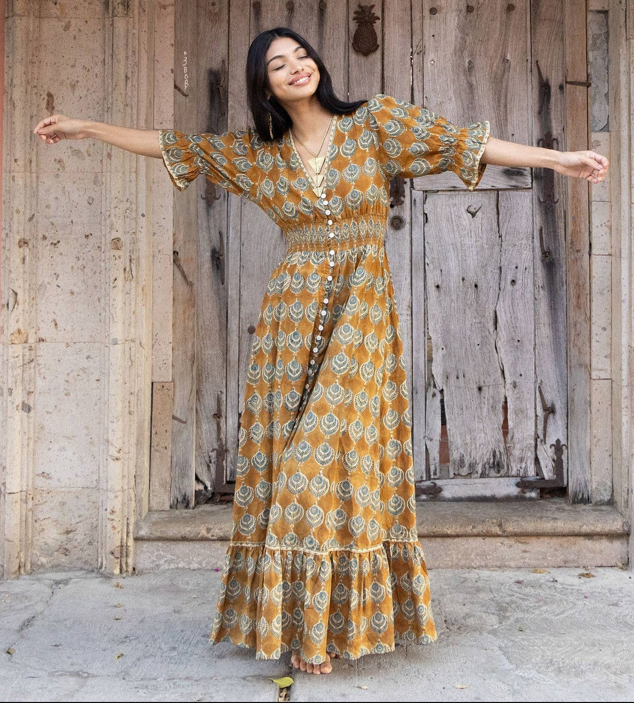Woman in a yellow dress standing under pink flowers with a wooden door and pink walls in the background