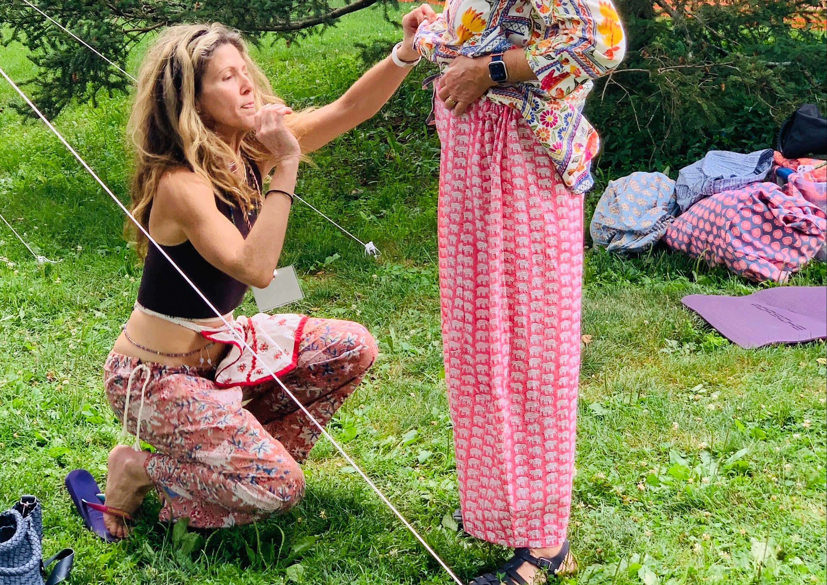 Two women in bohemian clothing standing on grass with a wooden fence and trees in the background.